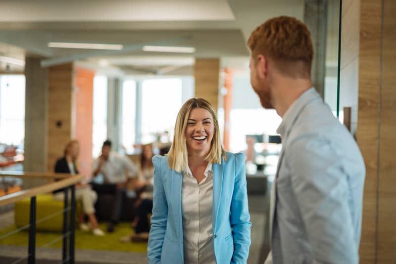 An office environment with a man talking to a woman who is smiling